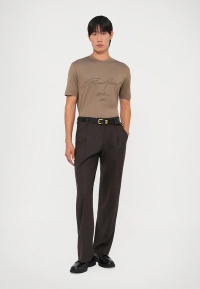Camiseta marrón de manga corta con un gráfico impreso, combinada con pantalones oscuros marrones y un cinturón negro con hebilla dorada, llevada con zapatos negros.