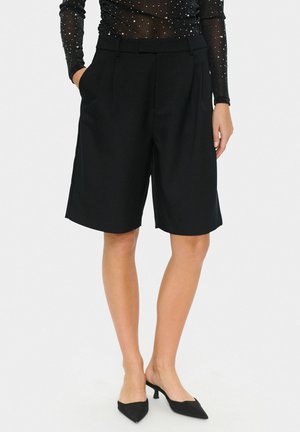 Schwarze maßgeschneiderte Shorts mit geradem Schnitt, gefälteltem Vorderteil und seitlichen Taschen. Getragen mit spitzen schwarzen High Heels und einem transparenten Langarmtop.