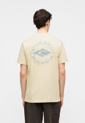 Νέος άνδρας με την πλάτη στραμμένη φορώντας ένα μπεζ T-shirt με μπλε λογότυπο «Rip Curl Surf Co Established 69» τυπωμένο στην πλάτη.