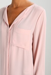 Blusa a maniche lunghe rosa chiaro realizzata in tessuto morbido. Presenta un collo a camicia e una tasca sul lato sinistro del petto. Texture liscia con vestibilità comoda.