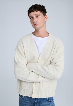 Cardigan lavorato a maglia di un beige chiaro con scollatura a V, polsini a coste e due tasche frontali. Presenta una chiusura con quattro bottoni e una consistenza morbida.