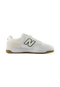 Witte sneaker met lage schuinstem, beige accenten, een gumzool, witte veters en een "N"-logo met zwarte rand aan de zijkant.