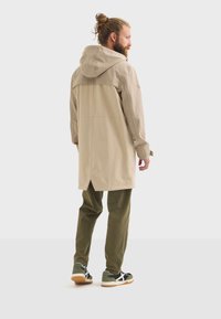Homme avec une barbe portant un long manteau beige, un pantalon vert olive et des baskets noires, se tenant de dos par rapport au spectateur sur un fond neutre.