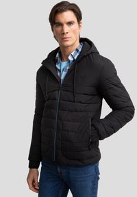 Giacca a vento nera con cappuccio, design trapuntato e accenti blu nella zip. Indossata sopra una camicia a quadri blu con colletto arrotondato.