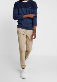 Navyblauer Strickpullover mit einem hellblauen horizontalen Streifen, kombiniert mit beigen Chinos und weißen Sneakers, die ein lässiges Outfit ergeben.