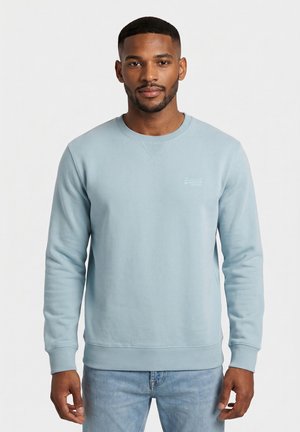 Joven con el pelo corto, vestido con sudadera azul claro de cuello redondo y vaqueros azul claro, posando frente a un fondo blanco liso.