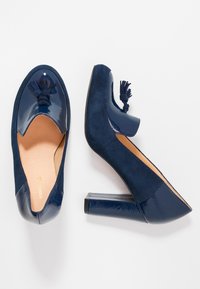 Tacchi alti blu navy con punta in vernice lucida e un esterno in morbido camoscio, caratterizzati da un dettaglio con nappina a forma di anello sulla tomaia e un tacco spesso.