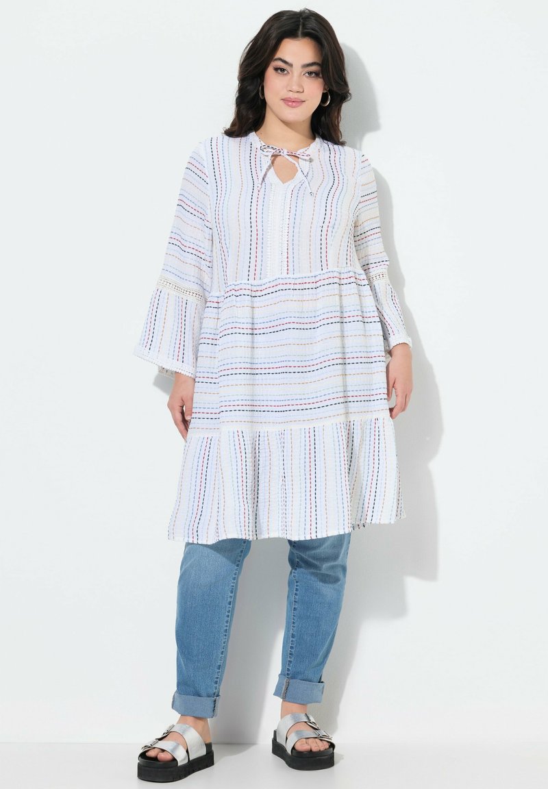 Weiß gestreiftes Kleid mit Glockenärmeln, Ausschnitt mit Schlüsselöffnung, geschichteter Saum, kombiniert mit hellblauen Jeans und silbernen Plateausandalen.