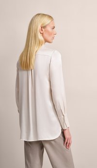 Light beige blouse in a smooth, satin fabric with long sleeves and a slightly curved hem, featuring a simplistic design and no visible patterns.