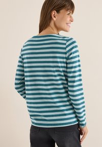 Kvinde, der smiler sidelæns iført en langærmet teal og lyseblå stribet skjorte samt sorte jeans foran en ensfarvet beige baggrund.