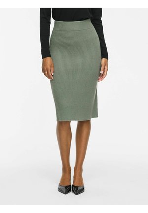 Donna che indossa una gonna a matita in maglia verde a coste aderente, una maglia nera a maniche lunghe e scarpe nere con punta appuntita, in piedi su uno sfondo semplice.