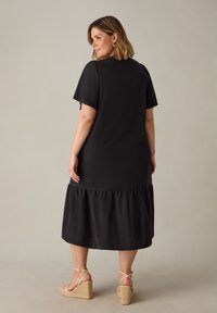 Schwarzes Midi-Kleid mit kurzen Ärmeln, gerüschtem Saum und weicher Textur. Kombiniert mit Riemchen-Wedge-Sandaletten mit geflochtenem Detail.