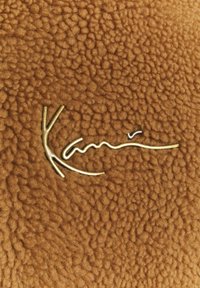 Nom de marque doré ressemblant à un fil "Karl" sur tissu brun texturé évoquant la polaire ou la laine.
