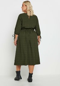 Vihreä ruudullinen mekko, jossa on väljä malli, kolme neljännesosaa hihoja, joustava vyötärö ja avainaukko takana. Yhdistetty mustiin nilkkabootseihin.