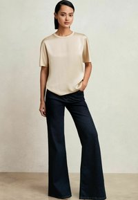 Femme debout, une main dans la poche, portant un haut beige en satin manches courtes et un jean large foncé sur un fond clair uni.