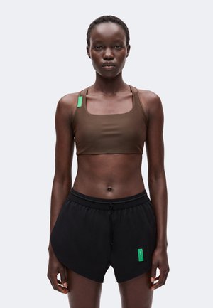 Kvinde med kort hår iført en brun croppet tanktop og sorte shorts med grønne mærker, står mod en ensartet lys baggrund.