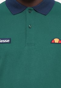 Zelená polo košeľa s námorníckym límcom, zapínaním na dva gombíky, vyšívaným logom na ľavej hrudi a kruhovým emblémom na pravej strane.