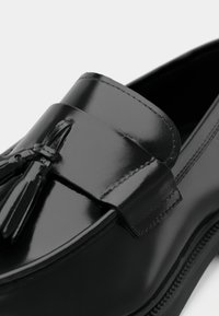 Black patent leather loafer with a sleek design, featuring a decorative tassel accent and contrasting stitching along the edge.
