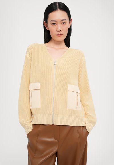 Cardigan com decote em V e fecho à frente em amarelo claro, feito de um tecido de malha com padrão texturizado e duas bolsas creme na parte da frente. Calças castanhas abaixo.