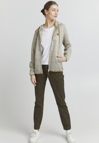 Veste à capuche avec un motif géométrique en beige et crème, dotée d'une fermeture éclair beige et d'un cordon de serrage, associée à un pantalon vert olive et des chaussures blanches.
