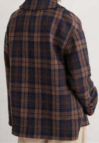 Tylny widok osoby w brązowo-granatowej w kratę wełnianej kurtce i jasnobeżowych spodniach na neutralnym tle.