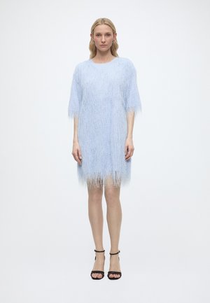 Woman standing wearing a light blue fuzzy knee-length dress with short sleeves and black ankle-strap high-heeled sandals.