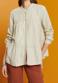 Blusa beige clara con cuello mao, botones al frente, cintura fruncida y mangas largas, combinada con pantalones de color óxido.