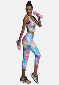 Kolorowy zestaw biustonosza sportowego i legginsów w wzór tie-dye. Trzyma różowe hantle, nosząc czarno-czerwone buty sportowe.