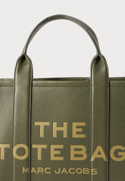 Olijfgroen leren toelatertas met een gestructureerd oppervlak, voorzien van grote gouden letters "THE TOTE BAG" en "MARC JACOBS" aan de voorkant. Handvatten zijn rechtop.