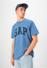 Tânăr îmbrăcat cu un tricou albastru GAP și blugi, cu o mână în buzunar, întorcându-se spre lateral pe un fundal alb simplu.