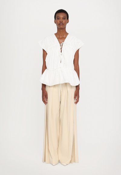 White cotton peplum top with a deep V-neck and lace-up front, paired with high-waisted beige wide-leg trousers. Smooth texture, flowing design.