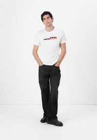 White cotton t-shirt with multicolor logo print; paired with black wide-leg jeans and black sneakers, standing against a white background.