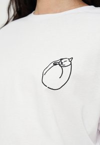 Chemise blanche avec un petit dessin au trait noir d’un chat recroquevillé sur la partie supérieure de la poitrine.