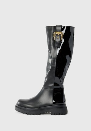 Bottes en caoutchouc noires, montantes jusqu'aux genoux, avec une finition laquée brillante, une semelle texturée et des détails en métal doré. Silhouette élégante et ajustée.