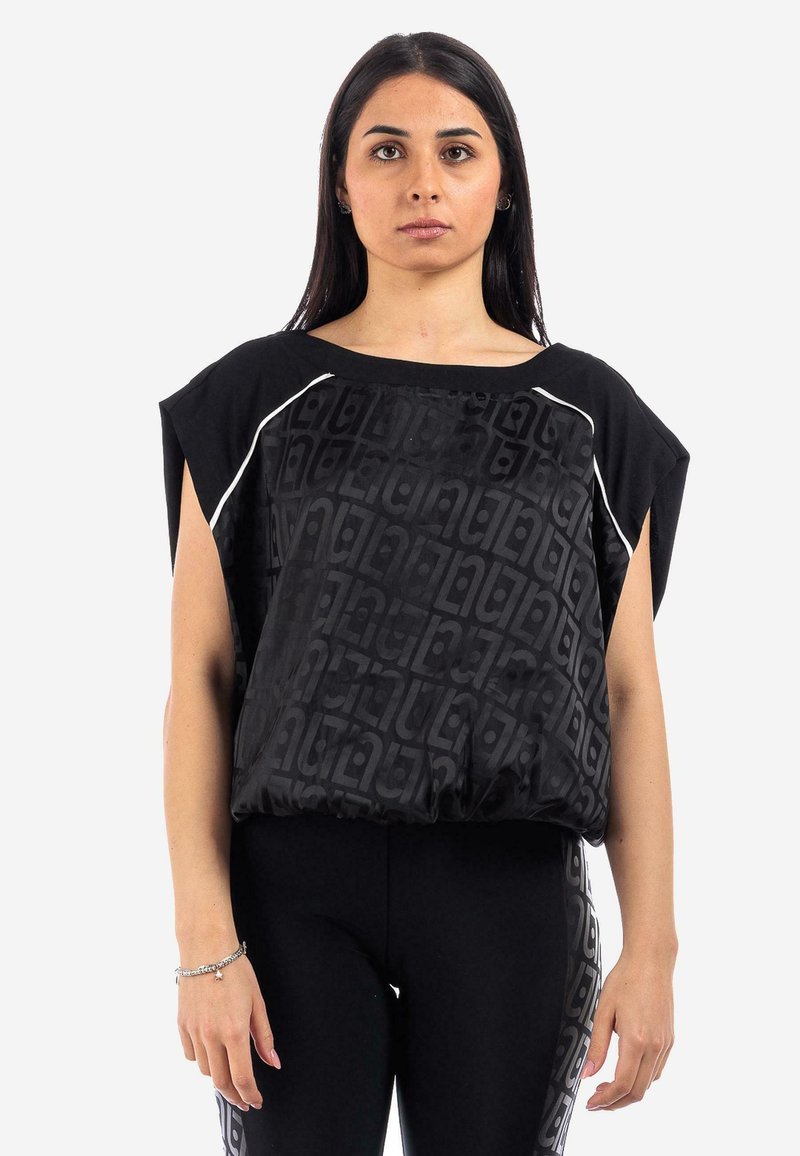 Top nero a maniche corte con un motivo testurizzato, dotato di accenti bianchi e una vestibilità larga. Indossato con leggings neri, sfondo neutro.