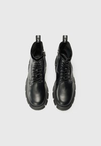 Zwarte leren enkelboots met vetersluiting aan de voorkant, gevoerde kraag, rits aan de zijkant en dikke rubberen zool. Voorzien van een aantrekband met branding.