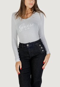 Top de manga larga color gris claro con el logo de "Guess" adornado con piedras de strass, combinado con pantalones de mezclilla oscuros de cintura alta con detalles de botones.