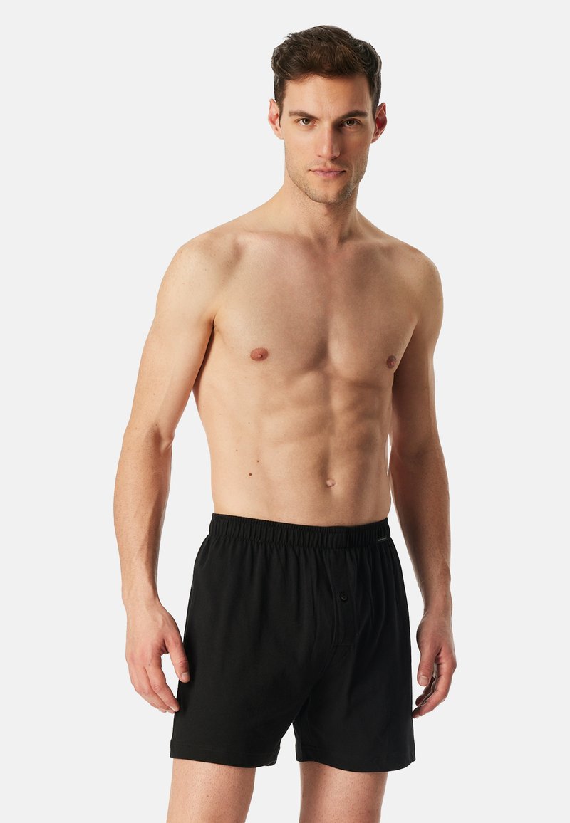 Shorts de boxeur noirs avec une taille élastique, dotés d'un bouton sur le devant et en tissu coton léger. Le modèle pose torse nu devant un fond uni.