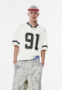 Hvid oversized trøje med sort numerisk tryk "91" og sorte striber på ærmerne, parret med mønstrede lyse cargo shorts.