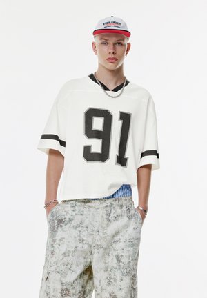 White oversized jersey with black numerical print "91" and black stripes on sleeves, paired with patterned light-colored cargo shorts.