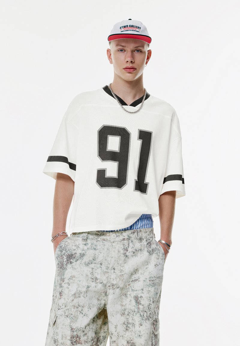 Hvid oversized trøje med sort numerisk tryk "91" og sorte striber på ærmerne, parret med mønstrede lyse cargo shorts.