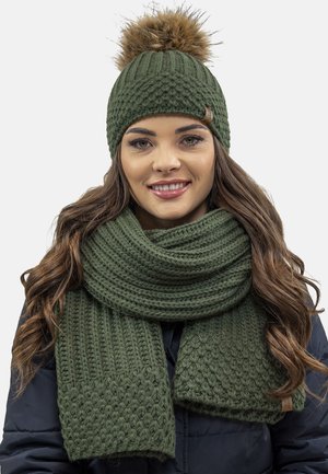 Green knitted beanie with a fur pom-pom and matching textured scarf. Both items feature a chunky, woven design.