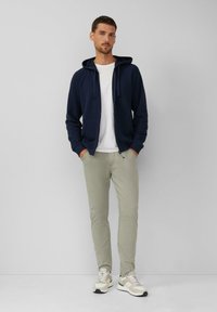 Felpa con cappuccio zip a navy sopra una maglietta bianca, abbinata a pantaloni a sigaretta verde chiaro e sneaker beige con accenti grigi.