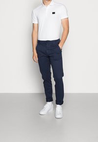 Vit pikétröja med korta ärmar och en liten svart logga, i kombination med marinblå chinos och vita sneakers. Släta tygtexturer synliga.