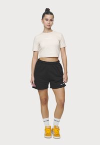 Béžové zkrácené tričko, černé šortky s kapsami, bílé logo. Model má na sobě bílé ponožky a oranžové tenisky. Jednoduchý design, ležérní styl.