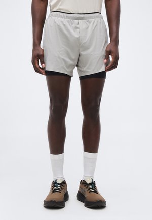 Moški, ki nosi svetlo sive večplastne tekaške kratke hlače, bele nogavice do gležnjev in rjave tekaške čevlje, stoji pred enobarvnim ozadjem.