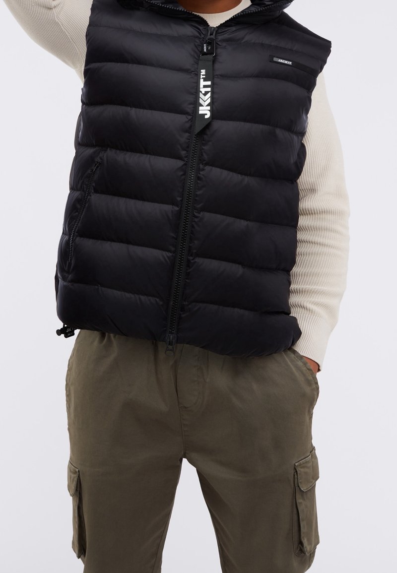 Veste sans manches matelassée noire avec fermeture éclair et logo sur un pull crème, associée à un pantalon cargo vert olive avec poches latérales.