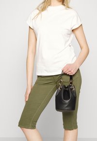 Žena v bílé košili s krátkými rukávy a olivově zelených zkrácených kalhotách, která drží malou černou koženou tašku ve tvaru kyblíku za ucho.