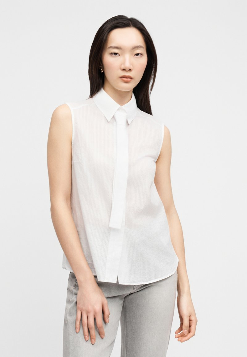 Femme asiatique aux longs cheveux noirs portant un chemisier blanc sans manches à col et un pantalon gris clair, debout devant un fond uni.