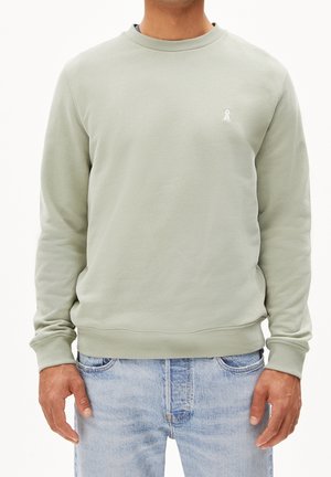 Mężczyzna ubrany w jasną, zieloną bluzę typu crewneck i jasnoniebieskie dżinsy, stojący ze swobodnie opuszczonymi rękami.